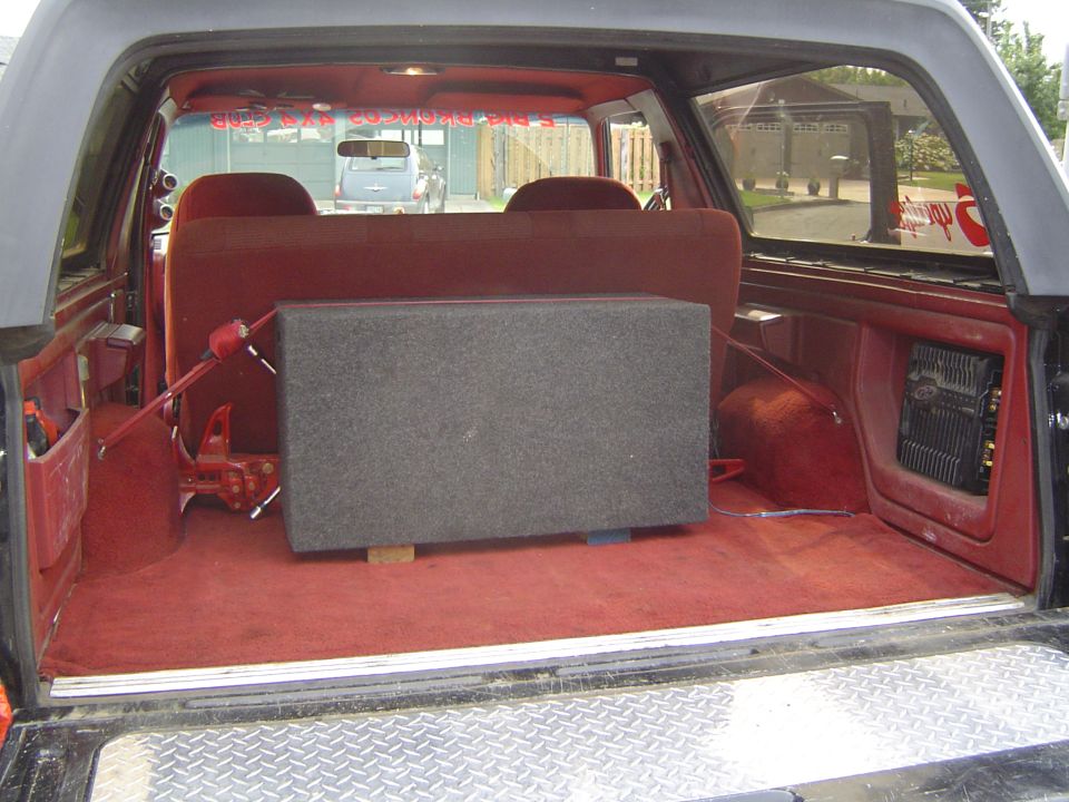 Red-carpeted SUV cargo area with gray subwoofer box and quarter-panel mounted amplifier