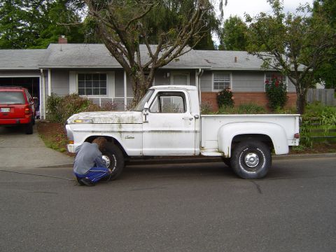 1972 Ford F-100 - A person works on the front tire of a white pickup truck parked on the street in front of a house.