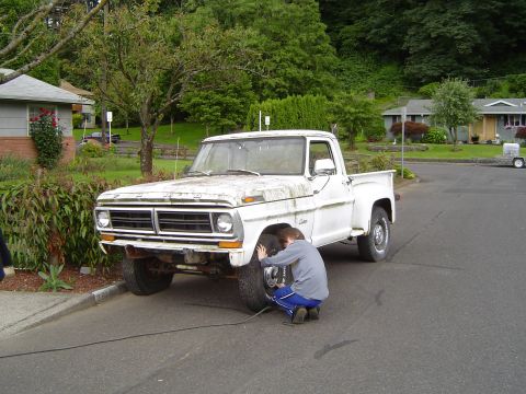1972 Ford F-100 - A person checks the front tire of a white 1972 Ford F-100 pickup truck parked on a residential street.