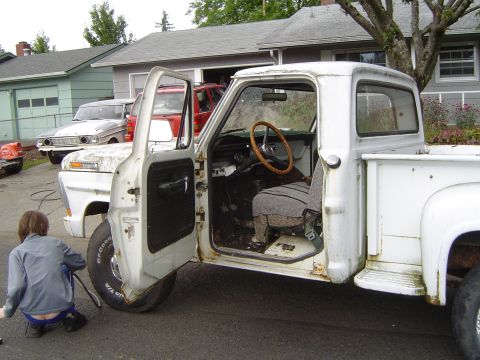 1972 Ford F-100 - A white 1967 Ford F-100 pickup truck sits in a driveway with its door open, as a person works on the front tire.