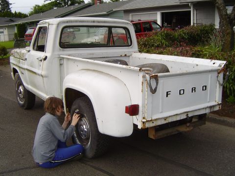 1972 Ford F-100 - White Ford pickup truck with a person attending to the rear tire. The tailgate clearly spells out 'FORD'.