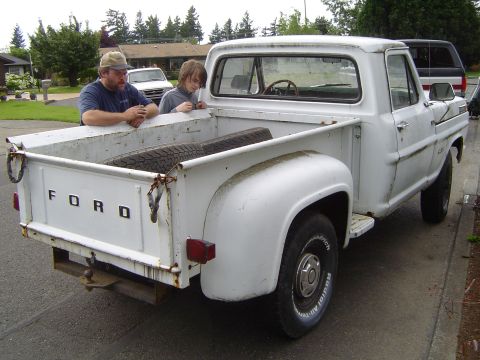 1972 Ford F-100 - White 1972 Ford F-100 pickup truck with two people in the bed, spare tire visible.