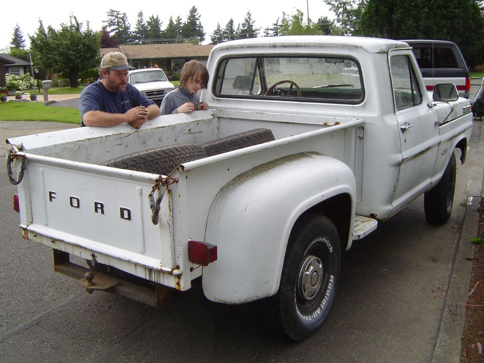 1972 Ford F-100 pickup truck, white, with two people looking into the bed. Spare tire is in the bed
