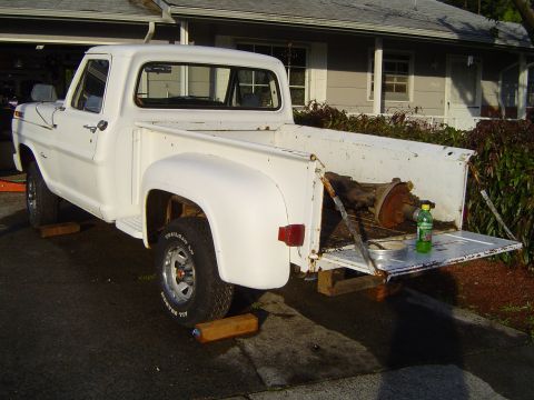 1972 Ford F-100 - This white 1972 Ford F-100 pickup truck is shown from the rear with its tailgate down, revealing a transmission stored in the bed.