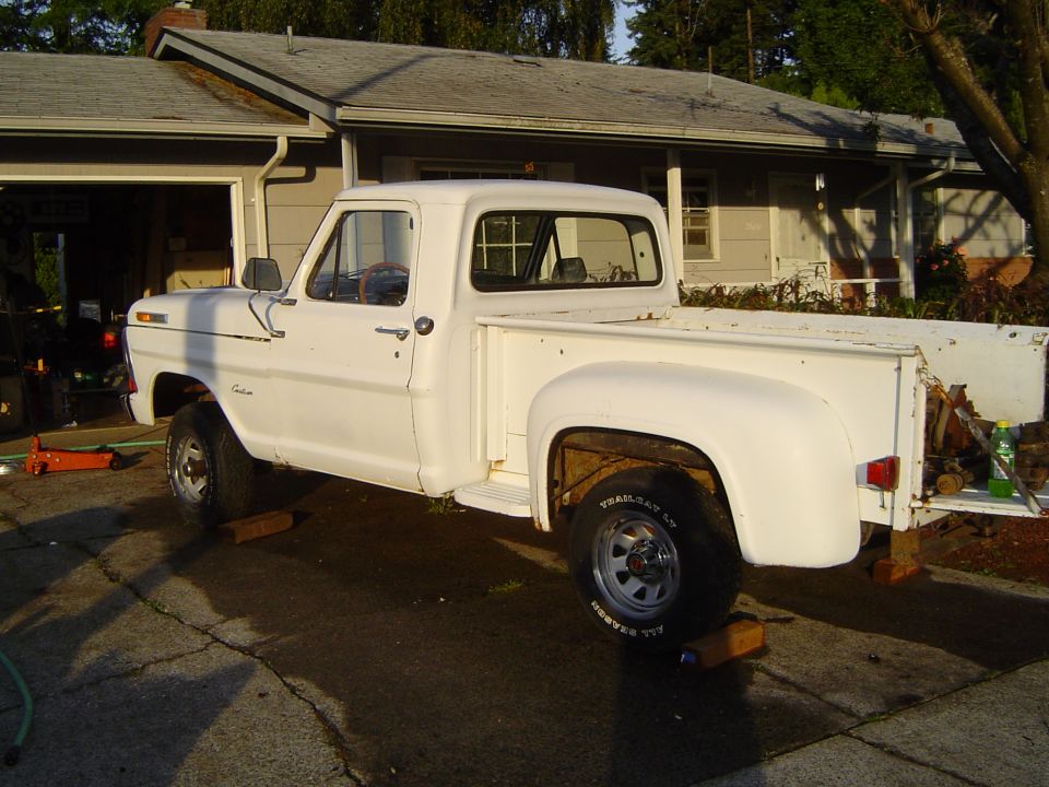White pickup truck, side view, raised on blocks, with visible rust on the rear wheel well