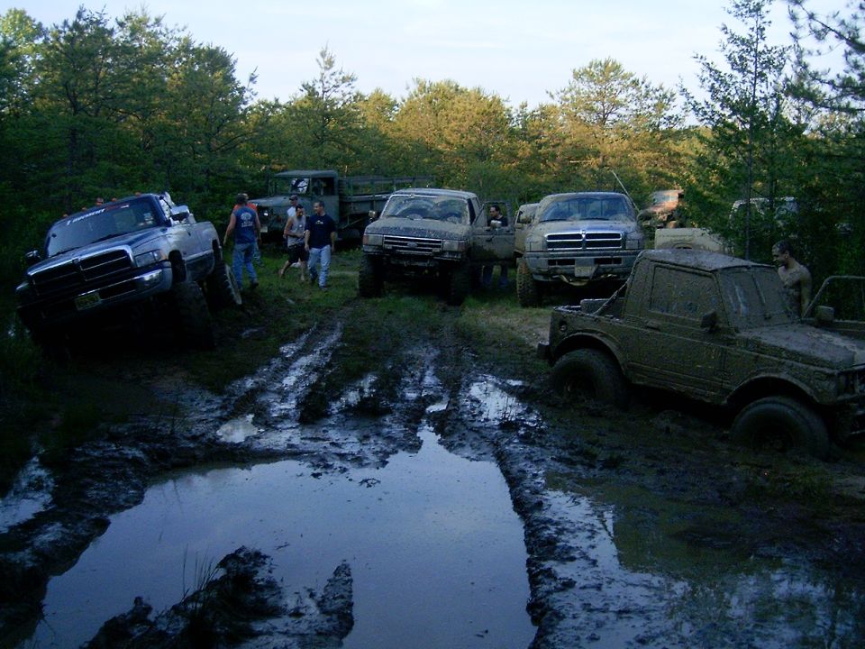 Lifted 4x4 trucks parked in a muddy forest trail with a large water-filled mud pit