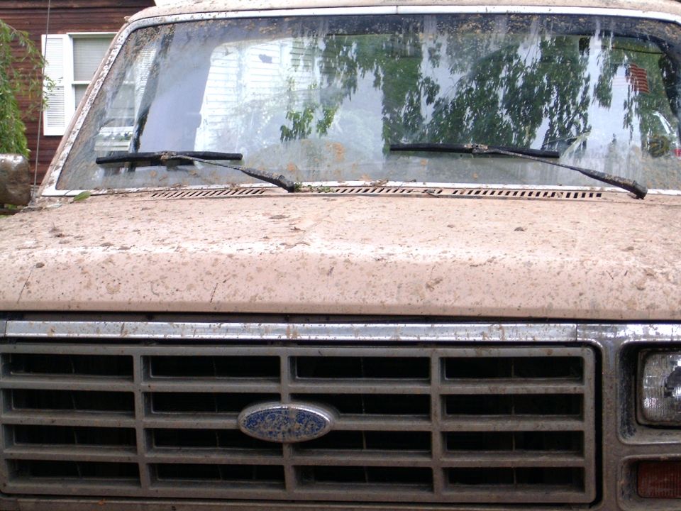 Front grille and cowl of a tan Ford truck splattered with dried mud, blue oval emblem