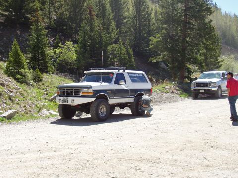 1979 Ford Bronco - A person works on the rear tire of a lifted Ford Bronco on a dirt road, with another truck in the background.