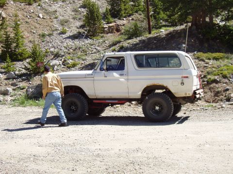1979 Ford Bronco - A man stands next to a white SUV with off-road modifications on a dirt road.