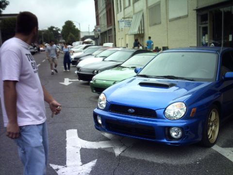 1995 Ford Probe - A man stands next to a line of cars parked on a street, with a blue Subaru Impreza WRX in the foreground.