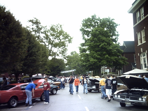 Red classic car with hood open, people milling around, brick building in background