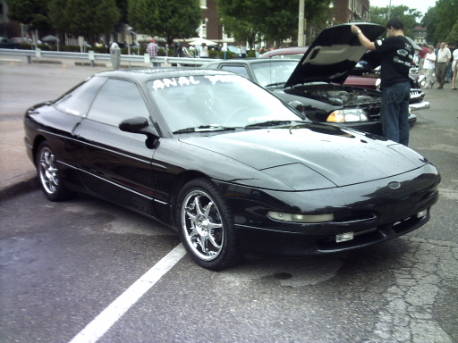 Black coupe with chrome wheels parked on asphalt. Another car's hood is open with a person looking inside