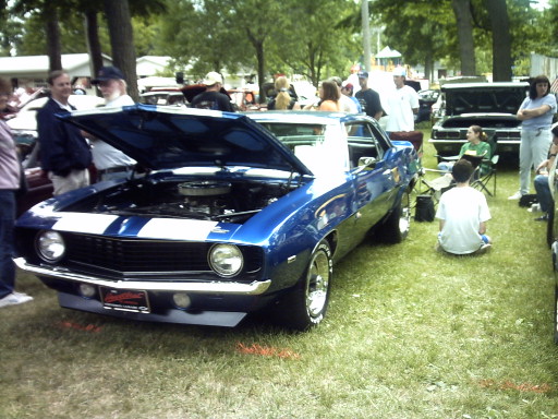 Blue classic car with white racing stripes, hood open, parked on grass at an outdoor event