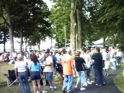 1995 Ford Probe - A large crowd of people are gathered outdoors under trees, with a paved walkway visible.