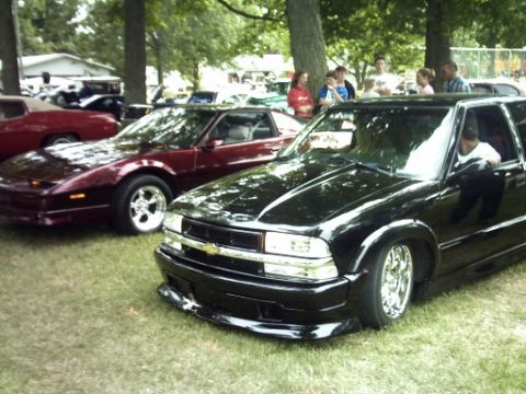 1995 Ford Probe - Several vehicles are parked on a grassy area under trees, with a group of people gathered in the background.