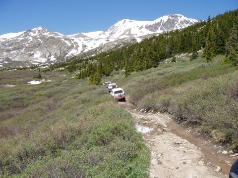 1979 Ford Bronco - A line of white SUVs navigates a challenging muddy track through a mountainous landscape under a clear blue sky.