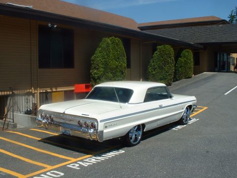 1999 Ford Crown Victoria - A white 1964 Chevrolet Impala convertible is parked in a lot with 'NO PARKING' painted on the asphalt.