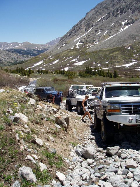 1979 Ford Bronco - A group of off-road vehicles are seen on a mountain trail, with a stream and rocky terrain.