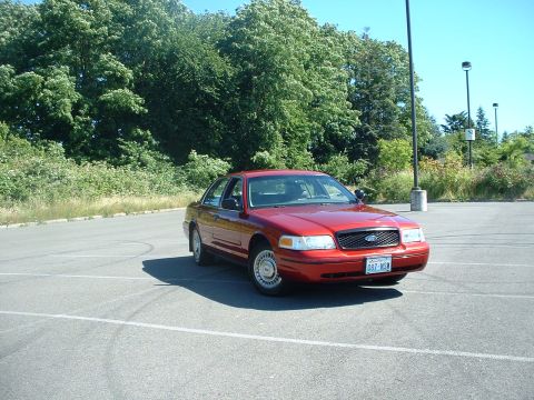 1999 Ford Crown Victoria - Red Ford Crown Victoria sedan parked in a paved lot with trees in the background.