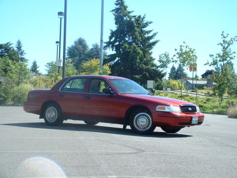 1999 Ford Crown Victoria - A red sedan is parked in an asphalt lot with trees and buildings in the background under a clear blue sky.
