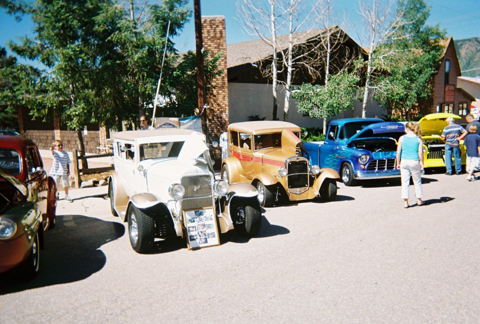 White 1930s Ford street rod at an outdoor car show with gold hot rod and blue pickup behind