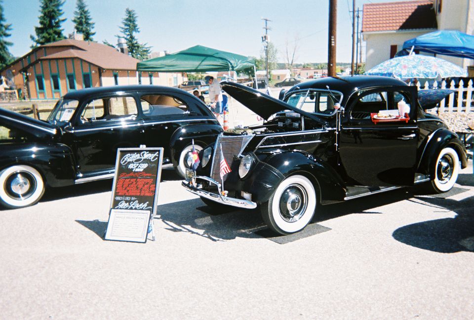Two black 1930s Ford coupes at an outdoor car show, hood up on the nearer car