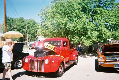 1972 Ford Ranchero - A bright red hot rod truck, modified with yellow flames and an open hood, is displayed at a car show alongside other vehicles.