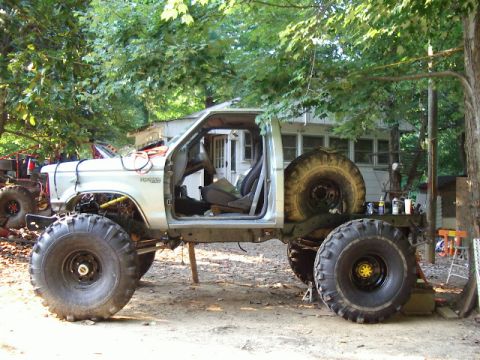 1985 Ford Bronco - This heavily modified pickup truck features oversized tires and a stripped-down body, with a second vehicle visible in the background.
