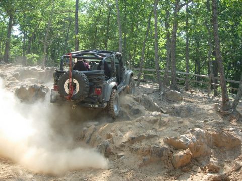 1995 Ford Bronco - A Jeep Wrangler navigates a dusty, rocky trail through a wooded area.
