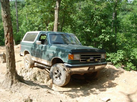 1995 Ford Bronco - Ford Bronco driving on a challenging off-road trail surrounded by trees.