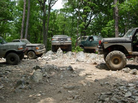 1995 Ford Bronco - Several off-road vehicles, including a 1995 Ford Bronco, traverse a challenging rocky path in a wooded area.