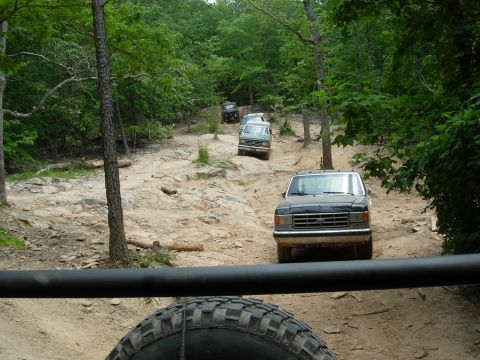 1995 Ford Bronco - Multiple Ford trucks navigate a challenging, rocky trail in a forest setting.