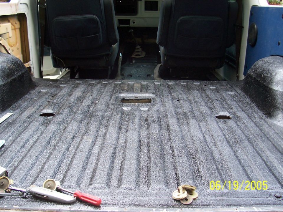 Vehicle interior floor with textured coating, showing two dark seats and tools