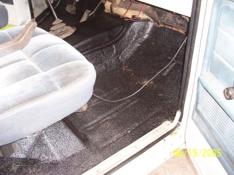 1979 Ford Bronco - The interior floor of a vehicle is covered in a black spray-on liner, with a portion of a light blue seat visible.