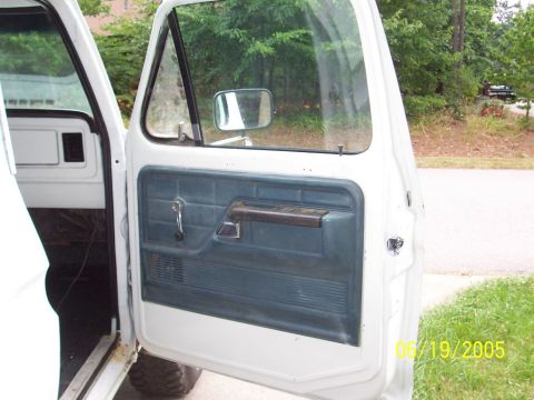 1979 Ford Bronco - Close-up of the driver's side door interior of a white Ford truck, blue door paneling and a chrome window crank.