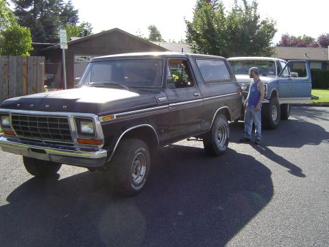 1979 Ford Bronco - A dark brown Ford Bronco is parked on a street next to a blue pickup truck with a person standing between them.