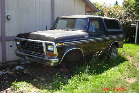 1979 Ford Bronco - A dark-colored Ford Bronco sits on jack stands with its front wheel removed, parked in a grassy area beside a structure.