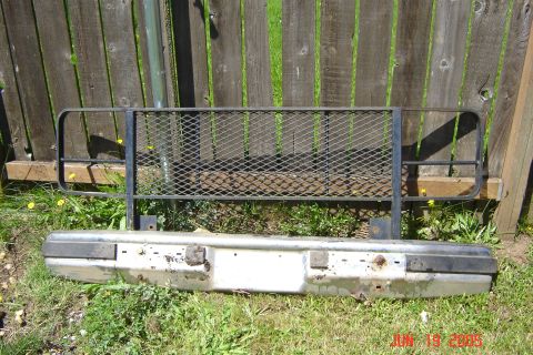 1979 Ford Bronco - Front bumper with a metal mesh brush guard attached, resting on grass in front of a wooden fence.
