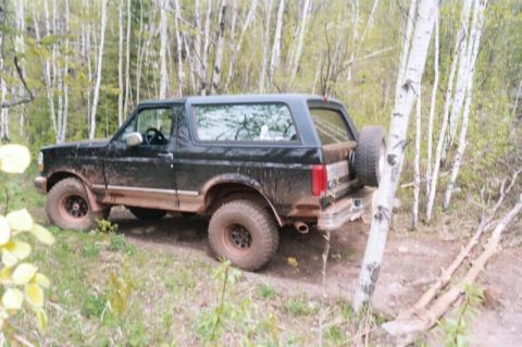 1996 Ford Bronco - Mud-covered 1996 Ford Bronco navigates a wooded trail with trees all around.