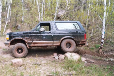 1996 Ford Bronco - Side profile of a black lifted full-size Bronco caked in dried mud along the rockers and lower body, sitting on black steel wheels with agg…