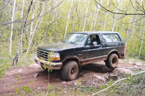 1996 Ford Bronco - Front three-quarter view of a black lifted full-size Bronco splattered with mud, flexing over a rocky dirt trail on mud-terrain tires and d…