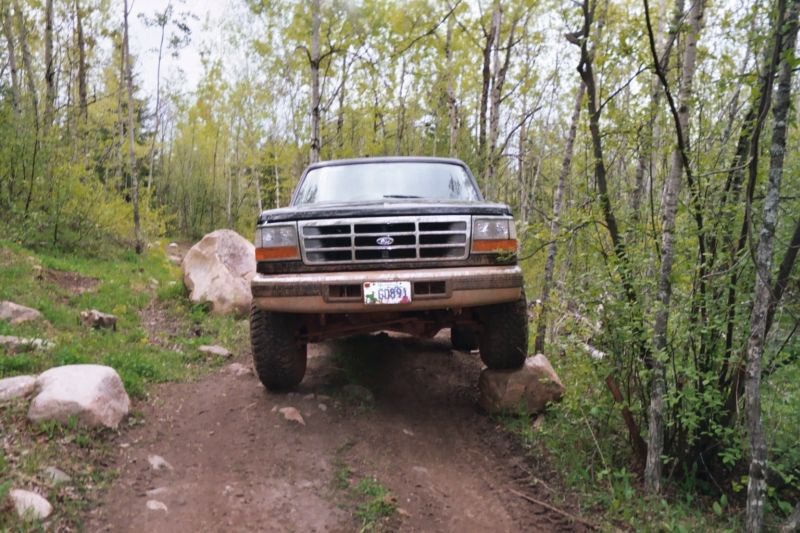 Muddy lifted Ford Bronco on a rocky wooded trail with bumper removed