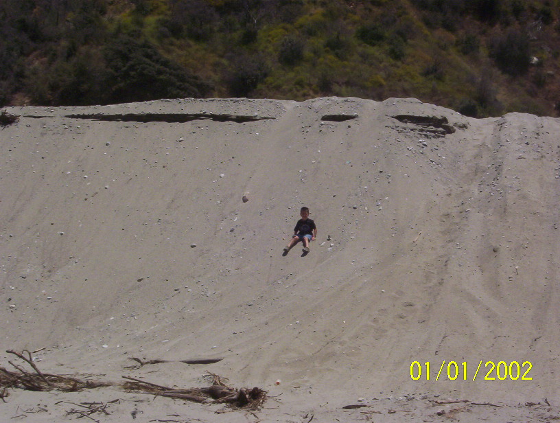 Child sitting on a sandy slope below a vegetated ridge, dated 01/01/2002.