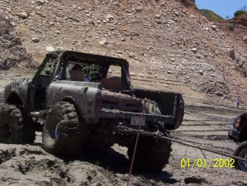 1985 Ford Bronco - A heavily mud-covered truck is stuck in a muddy area, with another vehicle assisting.
