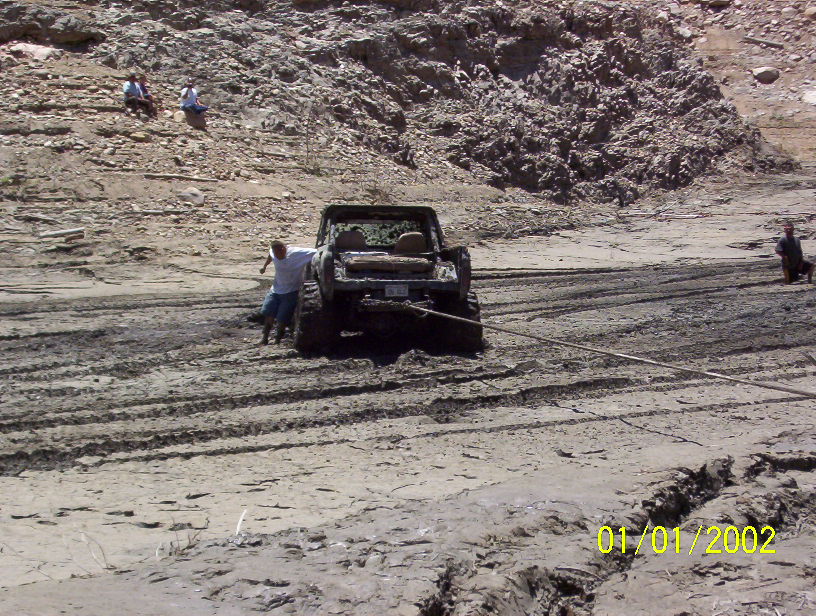 Mud-covered topless Bronco stuck in mud pit with tow rope attached, seen from the rear
