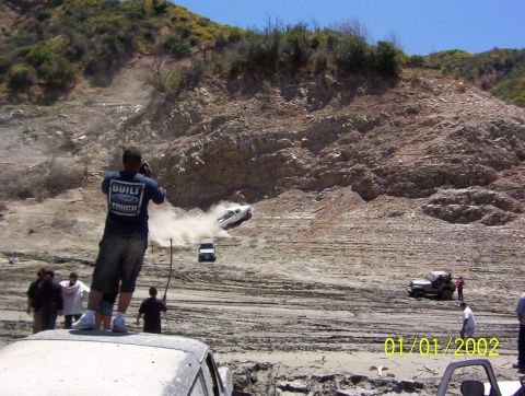 1985 Ford Bronco - Several off-road vehicles are seen in action on a dusty and muddy terrain, with onlookers observing the scene.