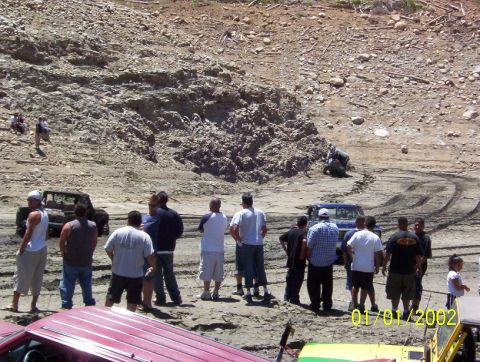 1985 Ford Bronco - People and off-road vehicles in a muddy area, with tire tracks visible on the ground.