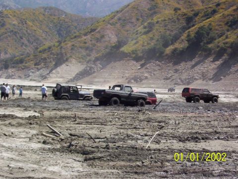 1985 Ford Bronco - Off-road vehicles navigate a challenging muddy terrain under a bright sky.