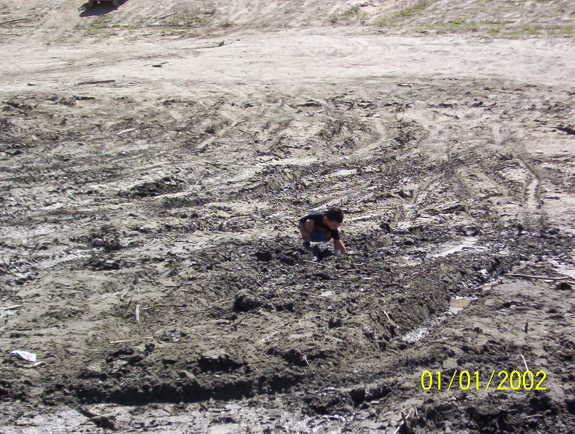 Person crouching in deep mud with tire tracks in a torn-up field, dated 01/01/2002