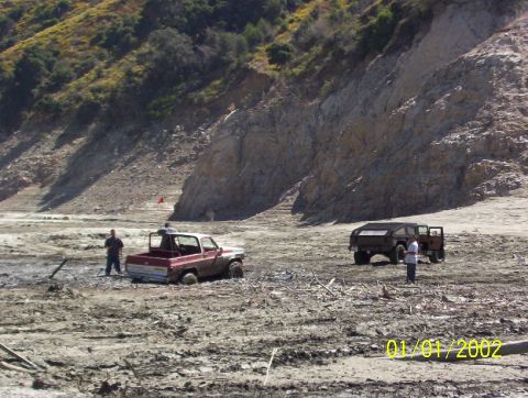 1985 Ford Bronco - Two off-road vehicles are stuck in a muddy, arid landscape with a rocky hillside in the background.